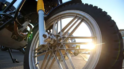 A close up of the front wheel of a motorbike. A low sun looming over the distant horizon shimmers through the spokes. 