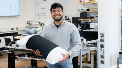PhD researcher Malindu Ehelagasthenna wearing a grey jumper and glasses, holding a pillow and smiling stood in a lab at Nottingham Trent University.