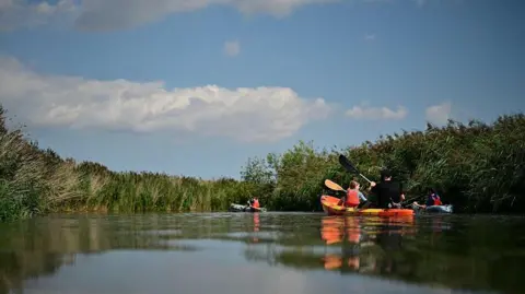 Three kayaks on a still river with high grass on its banks. The nearest kayak contains one adult in a hat and a child wearing a lifejacket. The sky is blue with white, fluffy clouds.