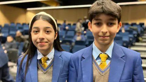 A boy and a girl stand next to each other in a large lecture theatre. they are wearing blue school uniform blazers with yellow ties.