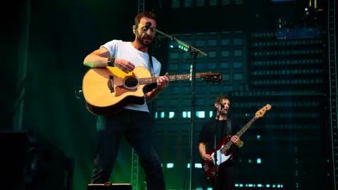 A Mulliner Captures Two men with brown hair and guitars on a stage with microphones, one in a white tshirt and the other in a black tshirt