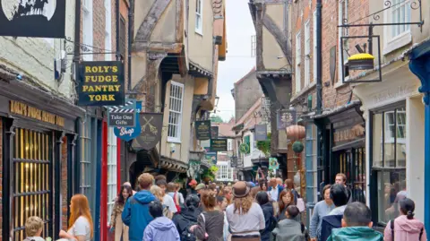 Getty Images View along The Shambles in York, a narrow medieval street with timber-framed buildings housing shops and cafes. The scene shows the busy pedestrian area that retains its characteristic overhanging upper stories and historic layout.