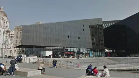 Image shows a pedestrianised area around Mann Island in Liverpool. A large black office block is in the background, with the Port of Liverpool building behind that.