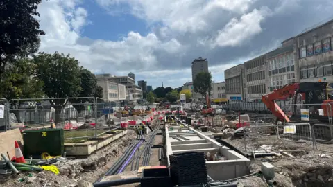 Plymouth Armada Way being dug up. There are diggers and soil. There are shops on either side. The image is looking down towards the sundial. There are trees to the left. 