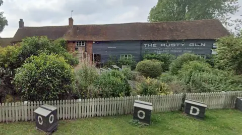 Google Single-storey grey pub building with "The Rusty Gun" on the back wall in white lettering with a grey background. There is a low wooden fence in the foreground with bushes behind it. There are bee hives in front of the fence.