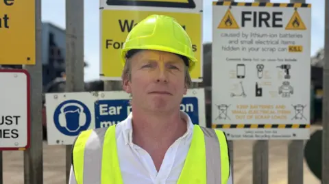 Man wearing yellow high-visibility jacket and helmet, stood in front of a lithium-ion battery fire warning sign.