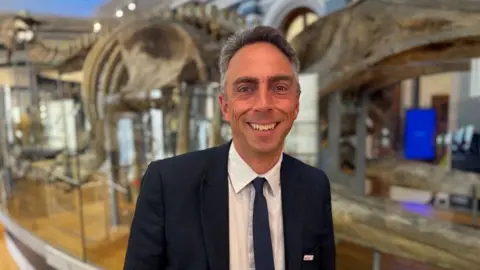 A man with grey hair and a navy suit and tie with a white shirt is smiling into the camera. The background is blurred but a large clear case has the skeleton of what appears to be a dinosaur in it.