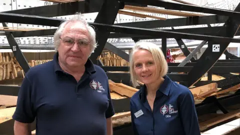 Darren Rozier/BBC Tim Kirk and Jacq Barnard both in dark blue Ships Company tops standing in front of frame with half built replica Sutton Hooship