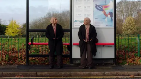 Muslim Women's Network UK Mariah is perched on a red seat at a bus stop in a winter coat, hijab and and has a bag on her shoulder. She is facing a double of herself, wearing the same thing and staring back at her.