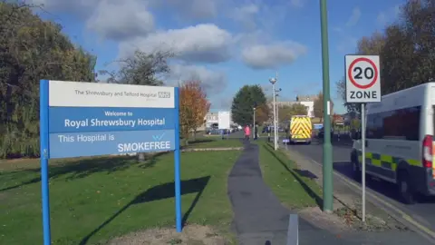There is a large blue and white sign at the corner of a road with the words "Royal Shrewsbury Hospital" on it. The sign is set on a grassed area with a path running alongside and trees in the background. There are two ambulances on the road that are heading towards white-coloured buildings in the far distance.