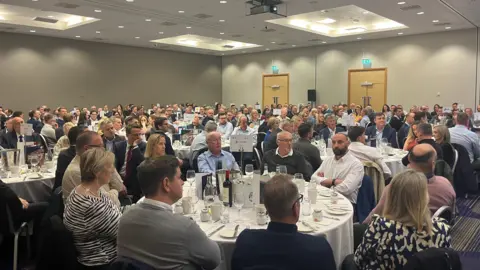 A packed room of business people sitting around round tables laid up for lunch. 