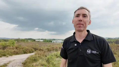 Chief executive of Devon Wildlife Trust, Nick Bruce-White is standing on heathland with cloudy skies overhead.