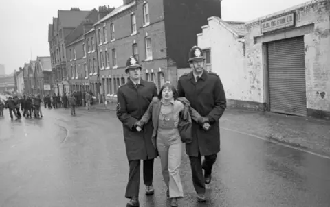 Sandra George Estate A young woman in dungarees in 1981 is being marched by two police officers in long coats and helmets up a street in front of a garage. A crowd can be seen in the background.