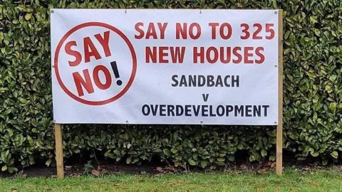 A large white banner alongside a hedge with red and black lettering which reads "Say no to 325 new houses - Sandbach v overdevelopment"