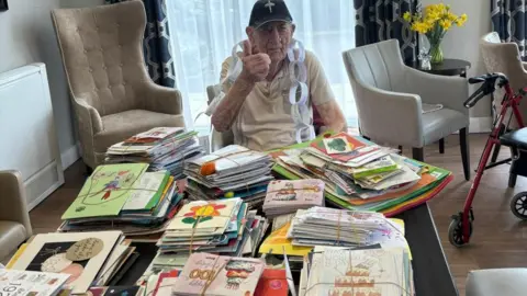 Stan Woolley, 100, wearing a black and green baseball cap with a military logo on the front and a beige polo neck, sits in front of a dining table laden with hundreds of birthday cards of all different colours. 