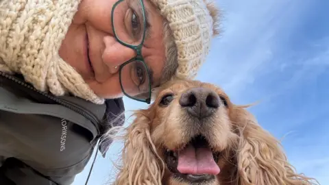 Supplied A woman in a woolly hat, matching cream scarf, glasses and a green coat is stood with her working cocker dog.