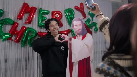 A man makes a heart sign with a cardboard cutout of a depiction of Jesus. A sign behind reads 'Merry Christmas'