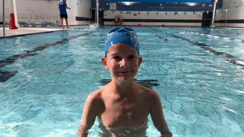 Bill, a young boy, smiles in a pool with a blue swimming cap on. 