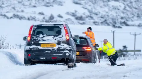 Michael Traill Two men wearing hi-vis coats attend to two cars parked at the side of a country road covered in snow