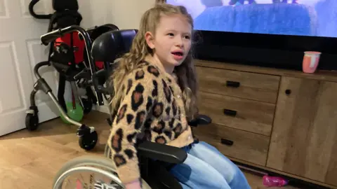 The young girl Alba is pictured in her current wheelchair in front of the TV in the family home