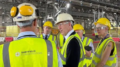 People stand around talking inside a warehouse. They wear helmets and neon yellow hi-vis. One is Dan Jarvis, who wears a suit under his vest.