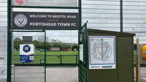 The image shows the entrance of Portishead Town FC grounds. There is a sign which reads "Welcome to Bristol Road, home of Portishead Town FC". The pitch is protected by green fencing. 