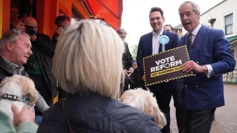 PA Media Nigel Farage dressed in a blue suit holds a Vote Reform sign towards a group of people sitting outdoors in a town centre. A candidate sporting a blue rosette is standing next to him.