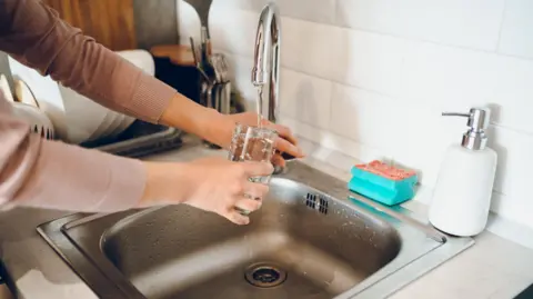 Close-up shot of a person at a sink holding a glass under a running tap. There is a sponge and a washing-up liquid container on the counter top.