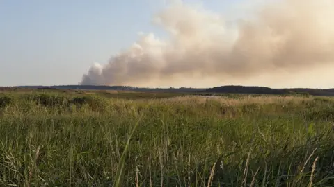 Rob Forsyth In a picture taken at ground level, grey smoke is drifting from the left to the right of the frame. There is grassland in the foreground.
