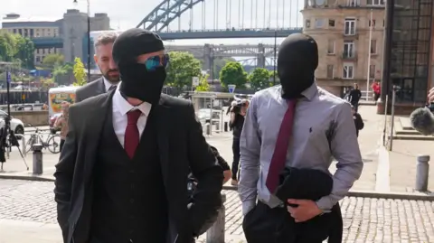 PA Media Graham and Carruthers walk into court. Both are wearing suits and have black hoods covering their faces