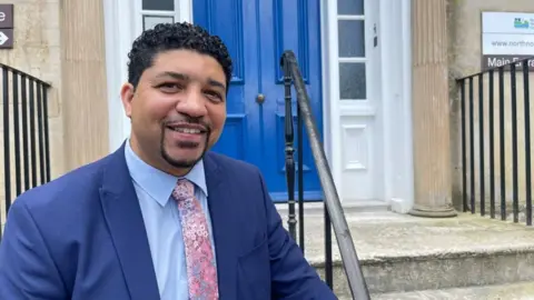 Glenroy Bell looking at the camera smiling, wearing a mid-blue suit, a light blue shirt and a pink floral tie. He is in front of same steps of a stone building leading to a blue door.