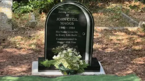 A black rounded top grave stone is inscribed with the details of a deceased person. There is a bouquet of pale flowers in front of it and it is in a dry grave yard area with small amounts of grass and leaves.