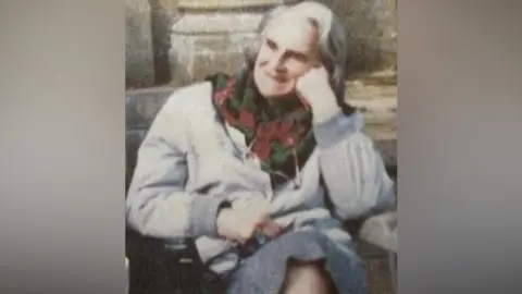 A grainy scan of a portrait photo of Mary Body sitting on a bench. She has grey shoulder-length hair and is wearing a light grey jacket and medium grey skirt along with a black, red and green floral scarf. She is also wearing glasses on a chain around her neck. She is looking off to the right of the camera, is smiling and has her left cheek propped up on her left knuckles. It can just about be made out that the wooden bench in directly in front of a stone building. 