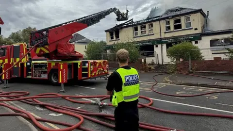 South Yorkshire Police A police officer stands in front of the burnt-out working men's club, with a fire engine parked outside.