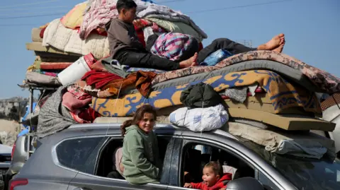 Reuters Palestinian children sit inside and on top of a car piled high with blankets, mattresses and other possessions which is queuing to go through a screening area on Salah al-Din Street, in the Netzarim corridor, in central Gaza (27 January 2025)