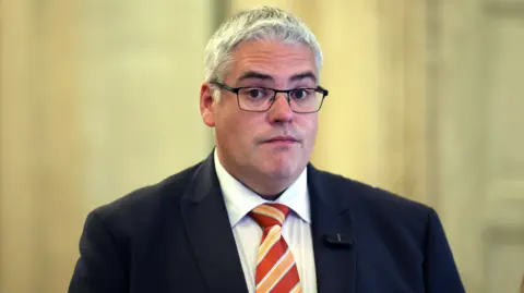 Gavin Robinson is wearing a navy suit with a white shirt and stripped tie. He wears glasses. The background is out of focus. 