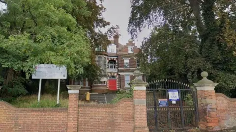 LDRS An elevated three story brick building with a red front door behind a wall and black wrought iron gates 