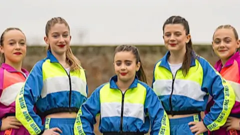 A line-up of four girls, each with hair scraped back in a ponytail, and wearing red lipstick and shellsuit-type outfits in various combinations of bright pinks, blues, purple and neon green.