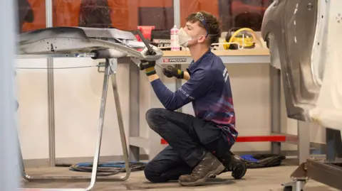 WorldSkills UK A man kneeling on the ground working on a car panel. He is wearing a navy long-sleeved polo shirt with red detailing, work trousers and brown boots. He has a pair of glasses on his head and is wearing a mask.