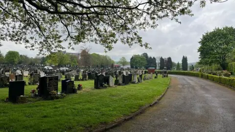 A photograph of a cemetery. On the left, there are dozens of gravestones on the grass and on the right is a path