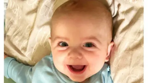 Baby Jensen-Lee Dougal smiling, a family photo which was shown to the jury at the start of the trial. He is wearing a blue top and his fine light hair. 