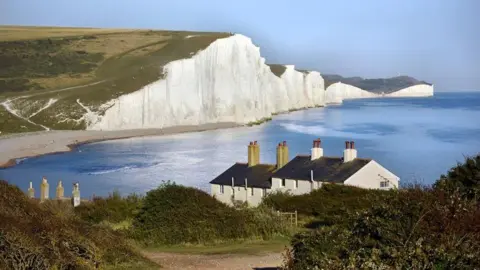 A view of the Seven Sisters cliffs and and Cuckmere Haven from cliffs near Seaford. The cliffs are white in the sunshine and the sea is blue. The cottages have white walls and chimneys and there is a gate leading to them.
