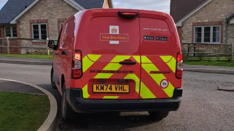 A Royal Mail Peugeot van parked by the side of the road in a residential neighbourhood. It is in red colours, with the Post Brenhinol logo on the back.