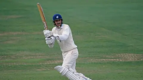 Getty Images A cricket player in a navy helmet holding a bat, having just struck the ball. He is wearing cricket whites and pads and about to run. He has a moustache and long brown hair.