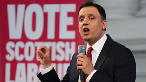 PA Media Anas Sarwar, in black suit, white shirt and red tie, is speaking into a microphone in front of a a red and white "Vote Scottish Labour" banner.