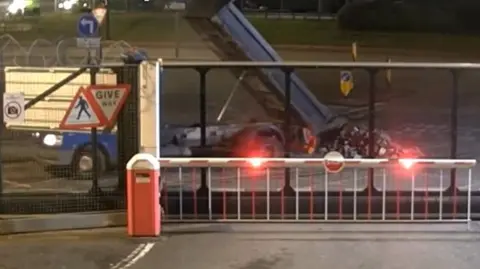 CPS A blue flat bed truck tips its back end up to pour a pile of rubble on to a road outside a security gate.