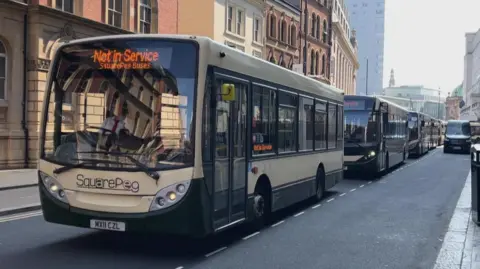Several single-decker buses which are green and cream and branded 'SquarePeg' in a row on a street in Leeds.