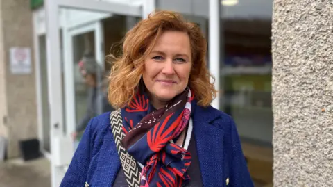 A woman with wavy brown hair, a colourful scarf and blue jacket is smiling at the camera, with doors behind her