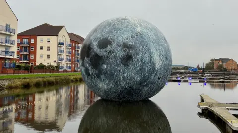 In the centre of the photo is a sphere printed to look like the moon. It sits in the middle of a body of water, surrounded by flats on the left, a pontoon on the right, and in the background, more buildings. The sky is grey.