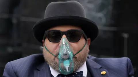 Nicholas Rossi, weaing a blue pin-stripe suit, bow tie and a black bowler hat. He is unshaven and is also wearing sunglasses and an oxygen mask.